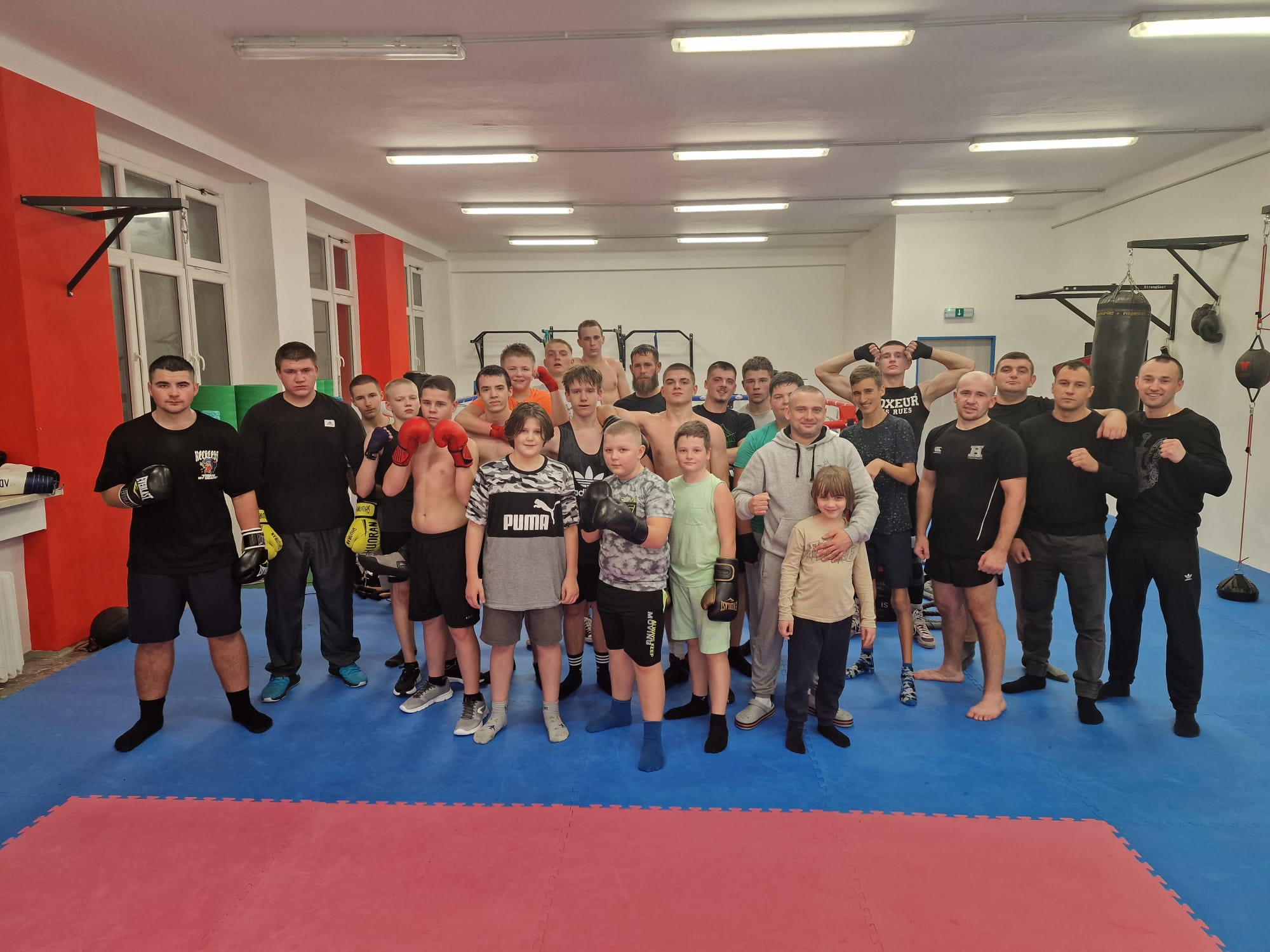 Skupina lidí různého věku v boxerské tělocvičně, připraveni na trénink, s boxerskými rukavicemi a sportovním vybavením.